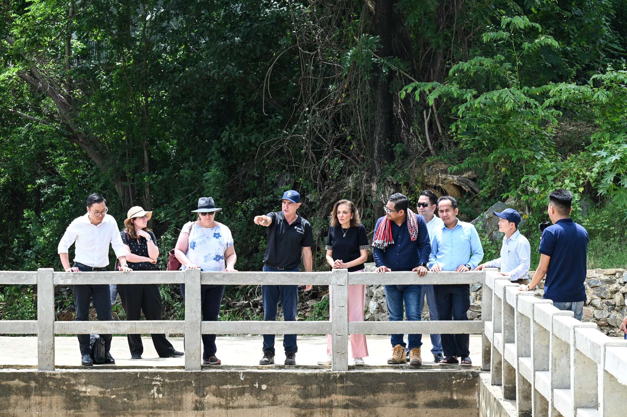 Group visits a water management site, standing on a bridge beside a tree-lined waterway.