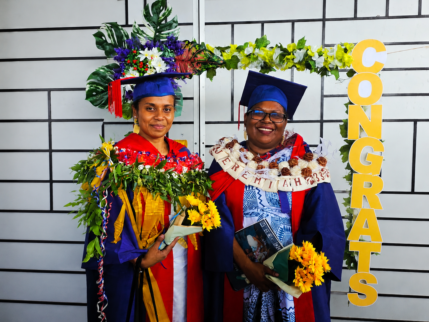 Two graduates in academic gowns holding flowers and posing at a graduation ceremony.