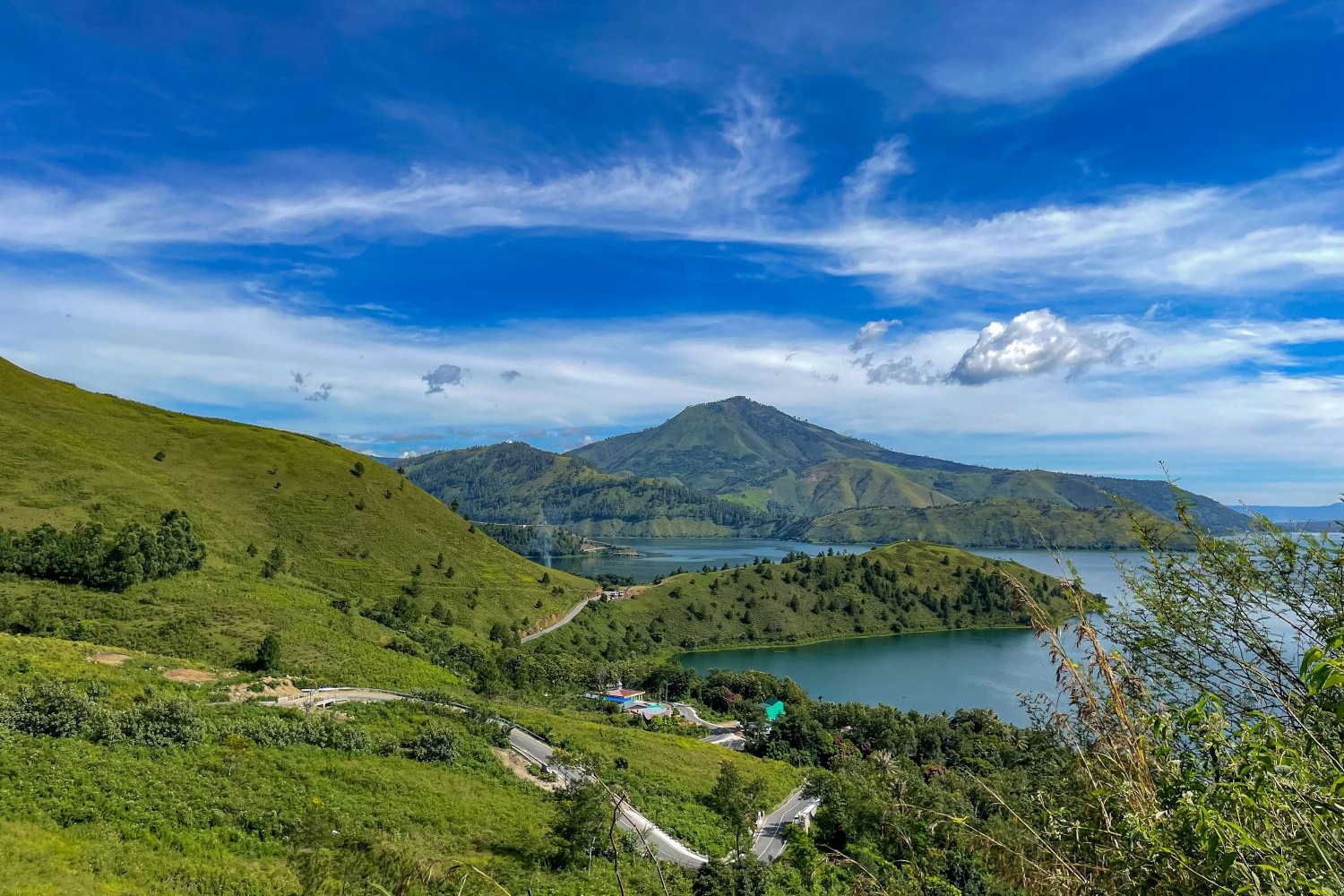 Green hills and a lake surrounded by mountains under a bright blue sky.