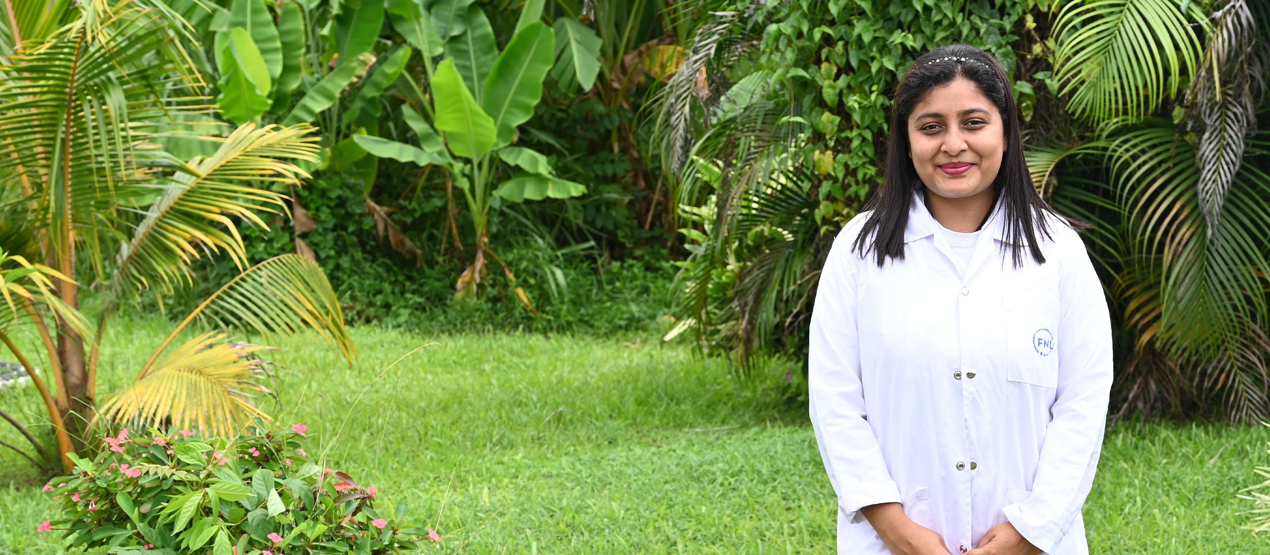 A woman wearing a white lab coat, standing on a green lawn with lush vegetation in the background, smiling at the camera. 