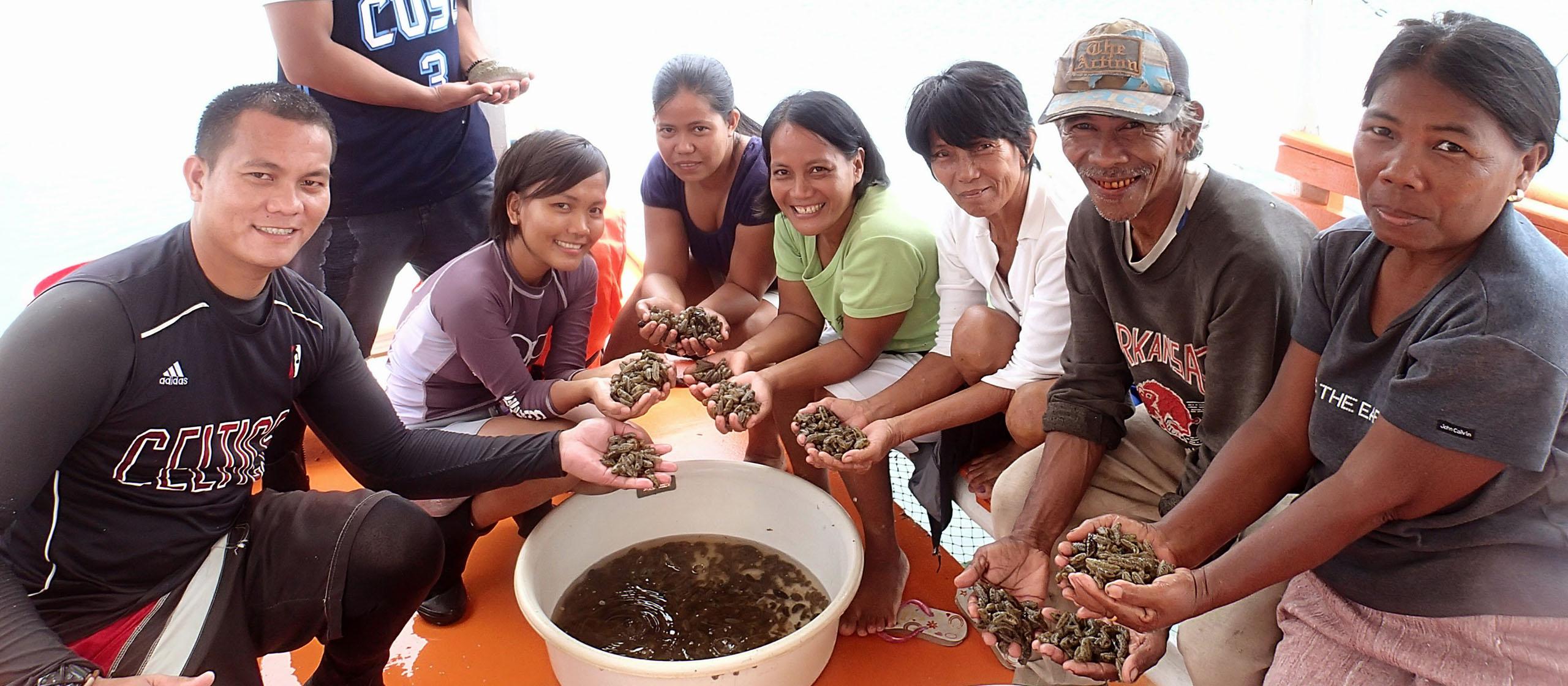 A group of people on a boat holding sea cucumbers, with a bowl of sea cucumbers in the foreground.