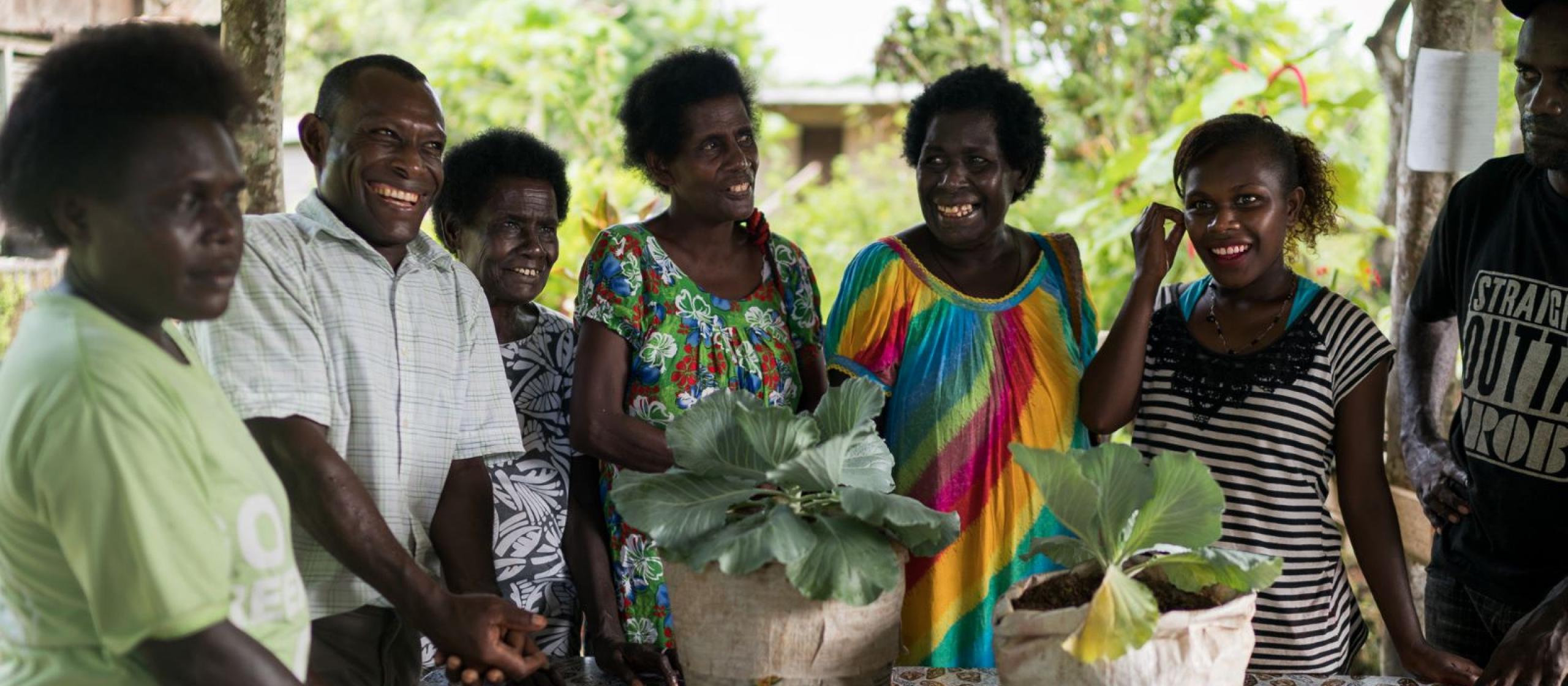 A group of people staning around a table containing two plants