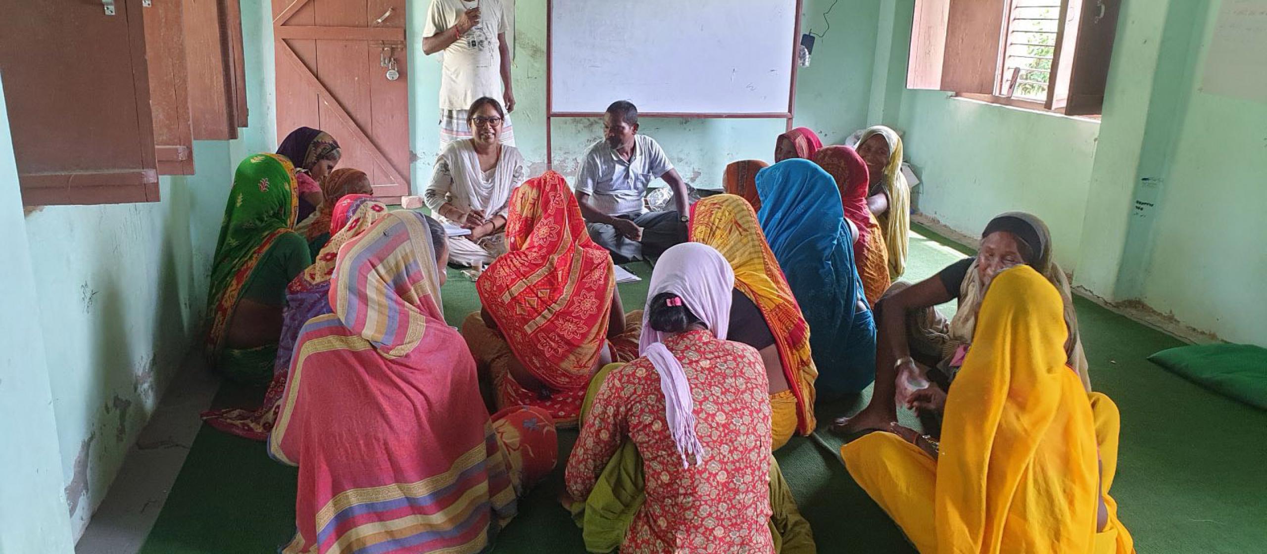 Group of women in colourful clothing seated in a circle indoors, listening to two facilitators, with a whiteboard behind them. 