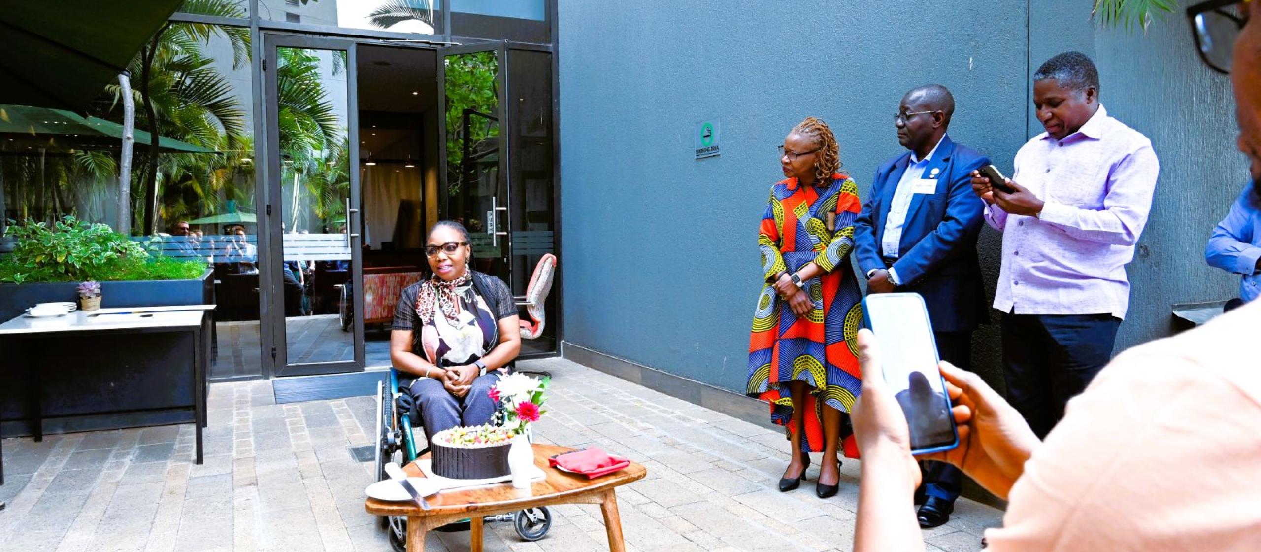 A small outdoor gathering where a speaker addresses attendees near a table with a cake, outside a modern building with greenery.