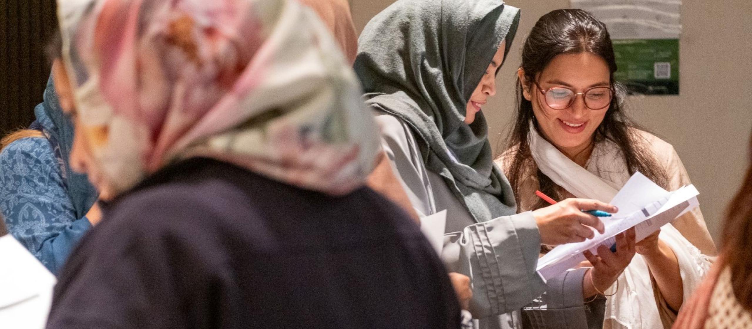 Women standing together indoors, reviewing documents and discussing notes during a collaborative workshop.