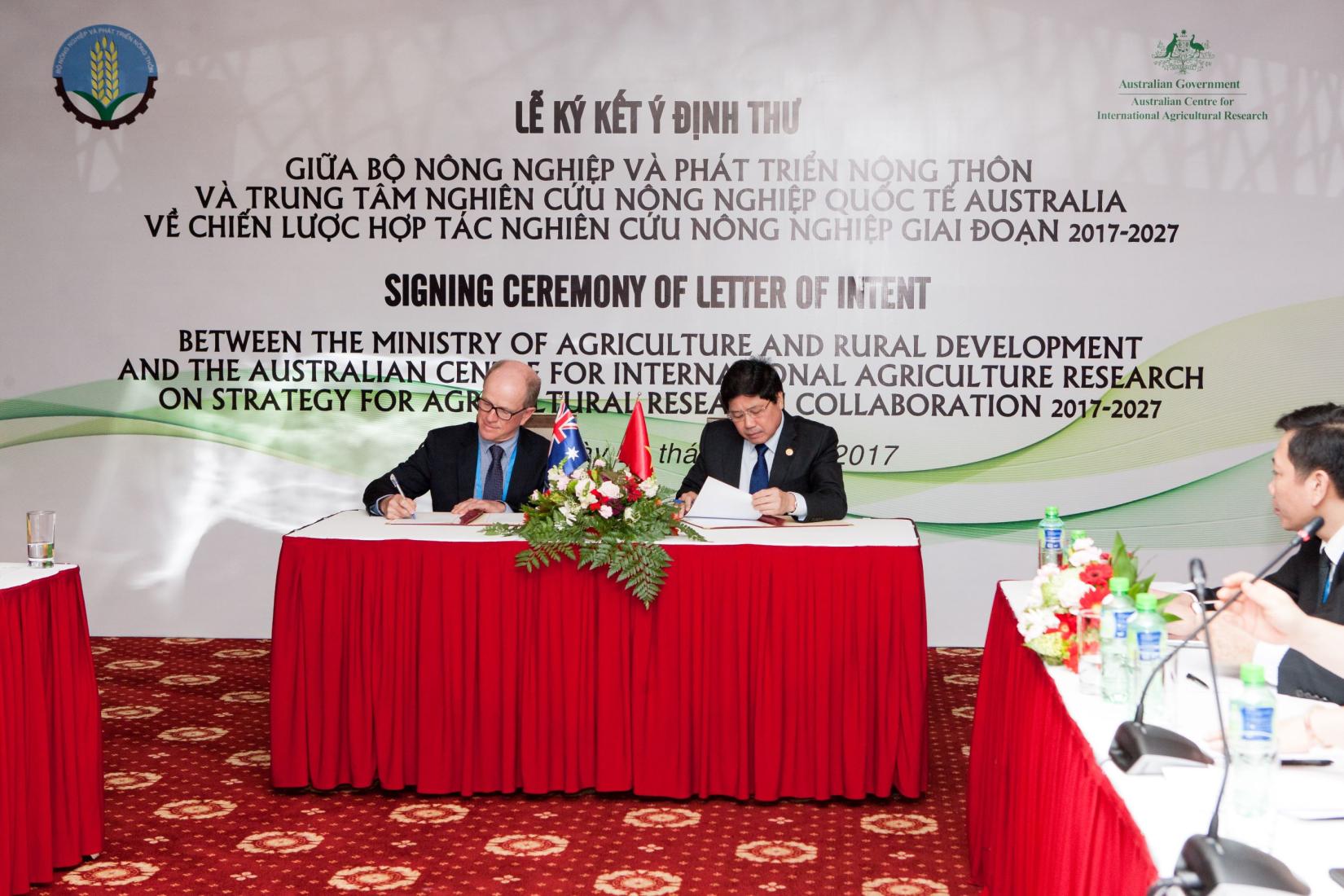 Two men sitting a an official table with a red tablecloth signing documents