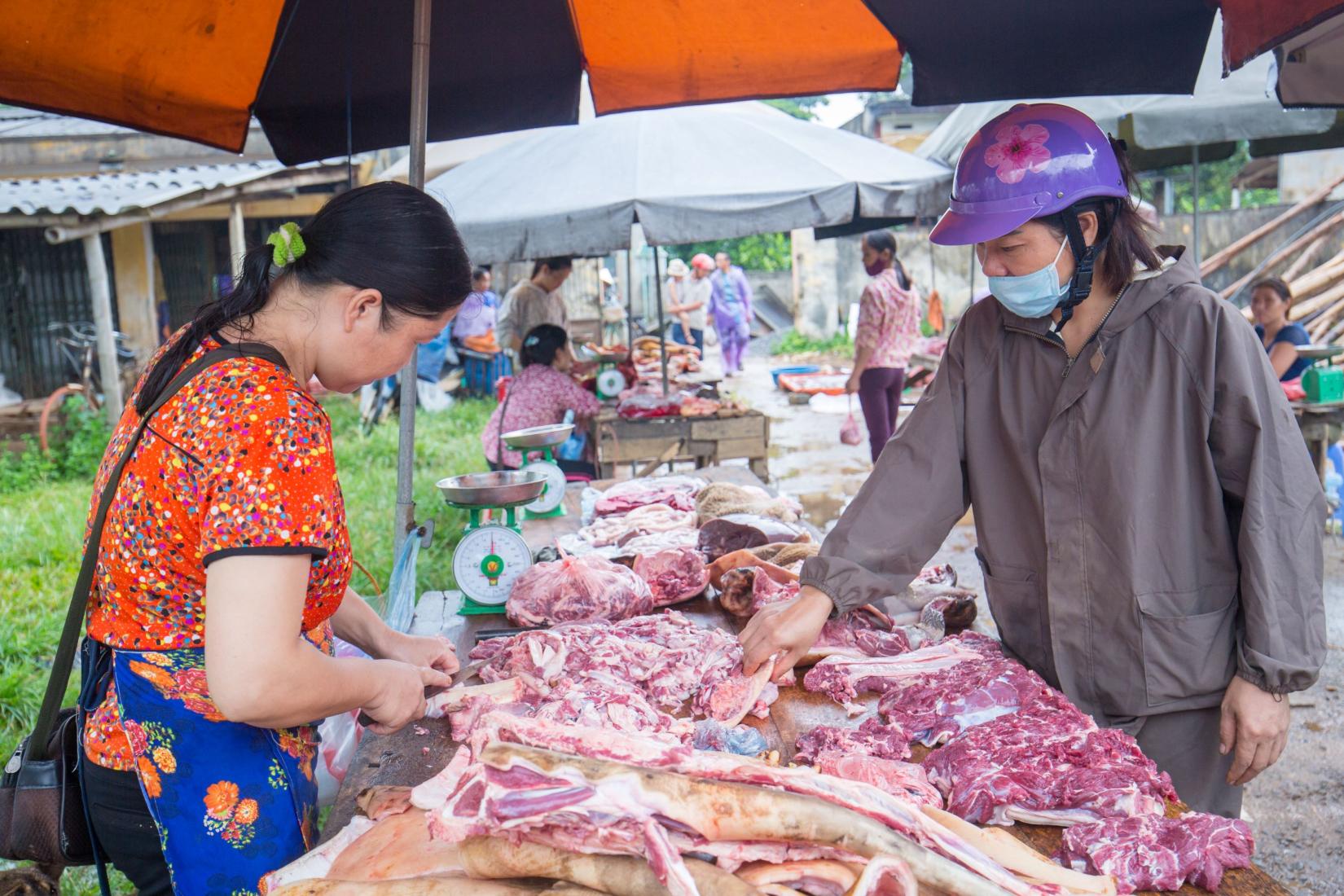 Vendor at market stall cutting meat with another person inspecting meat