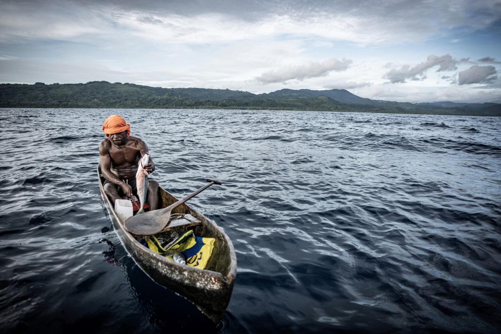 A man sits in a wooden canoe on the water holding a freshly caught fish