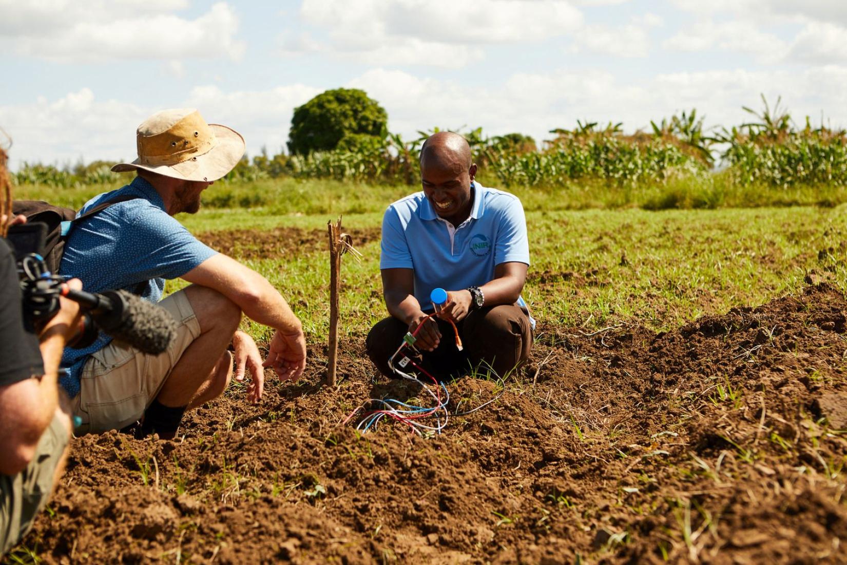 Two men crouch in a field, examining soil and monitoring equipment placed in the ground. 