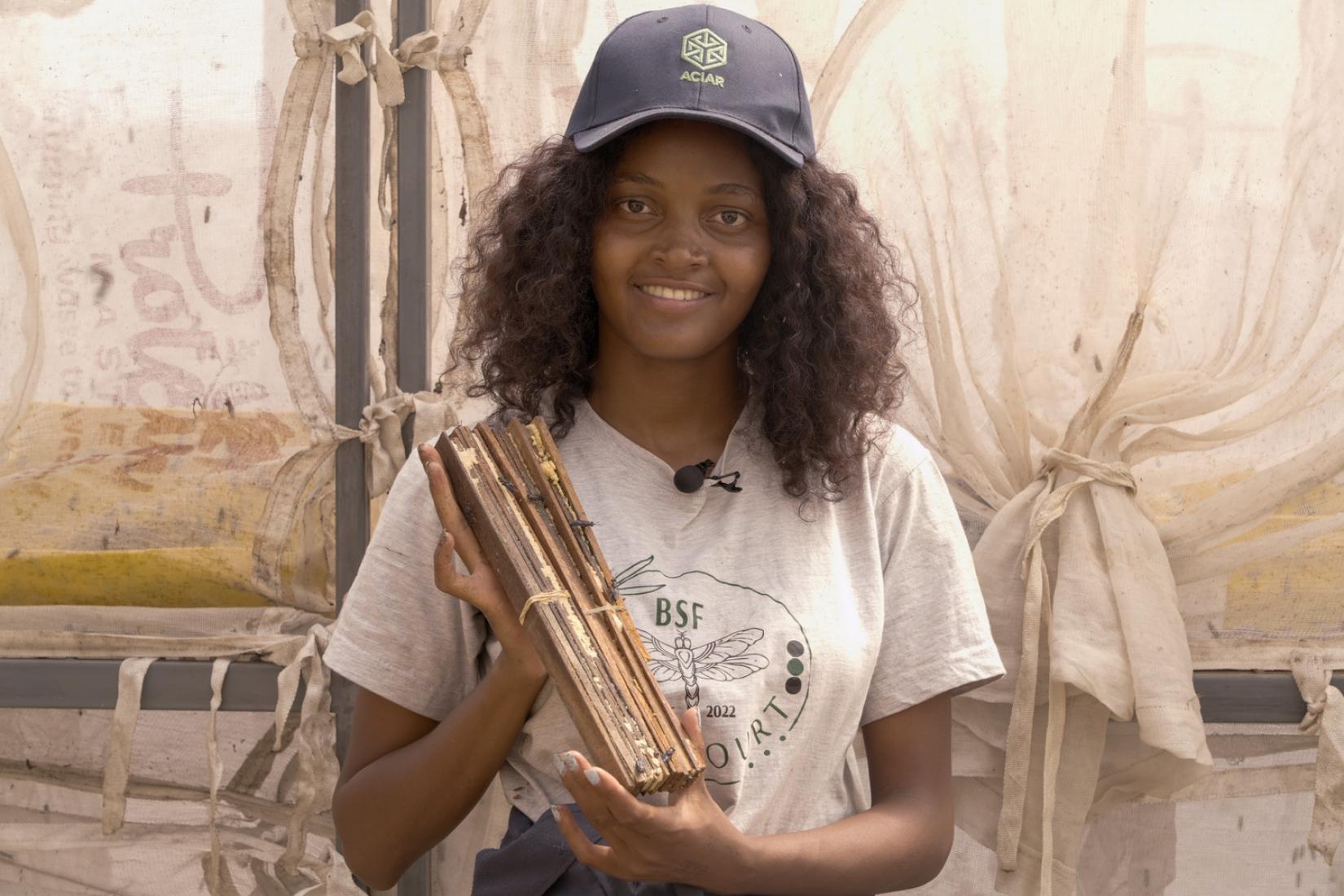 A woman wearing an ACIAR cap smiles at the camera while holding a bundle of stacked wooden boards.