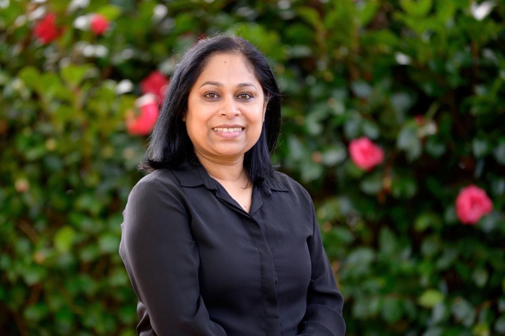 Portrait of Dipika in a dark-coloured shirt smiling at the camera with green foliage in the background. 