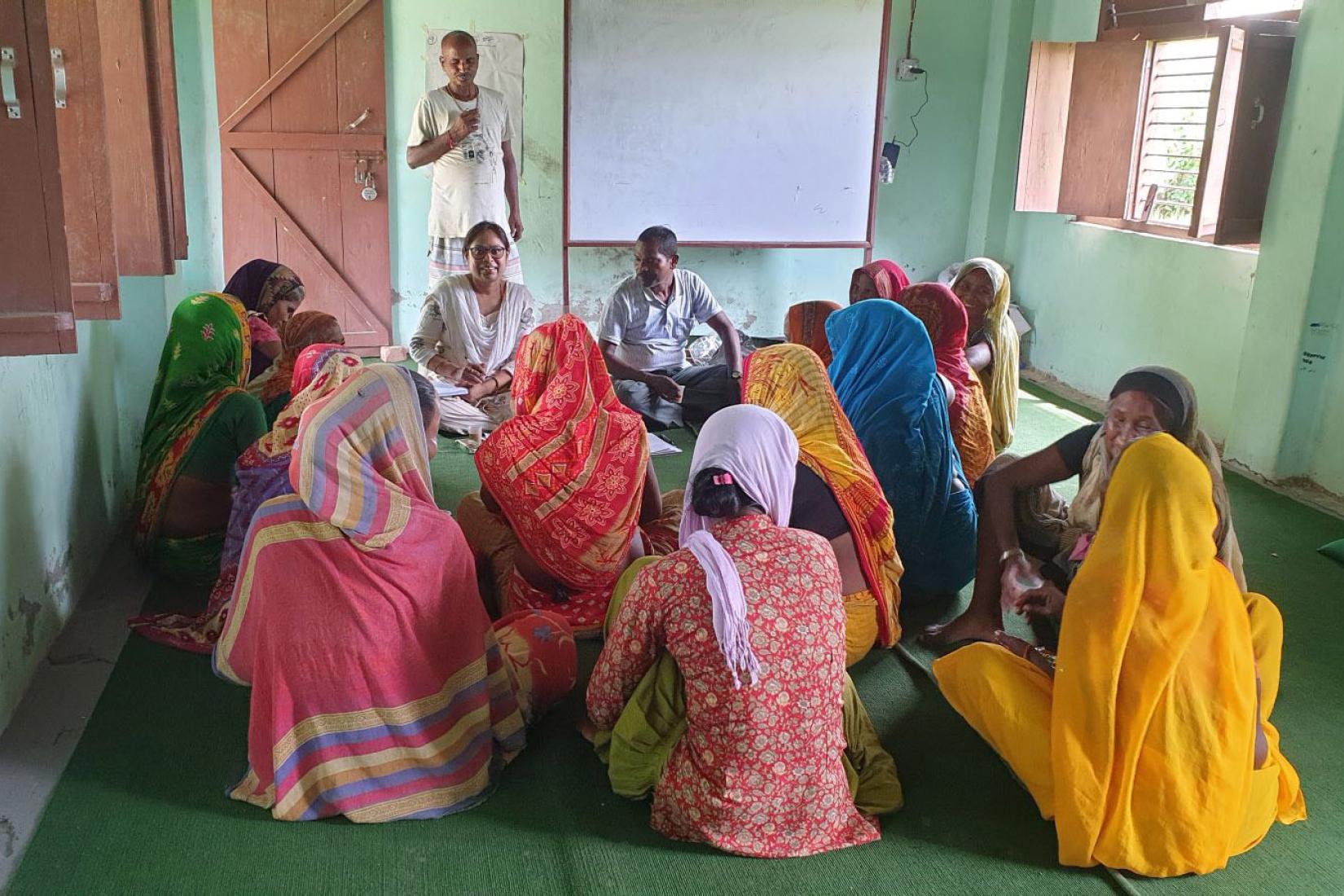 Group of women in colourful clothing seated in a circle indoors, listening to two facilitators, with a whiteboard behind them. 