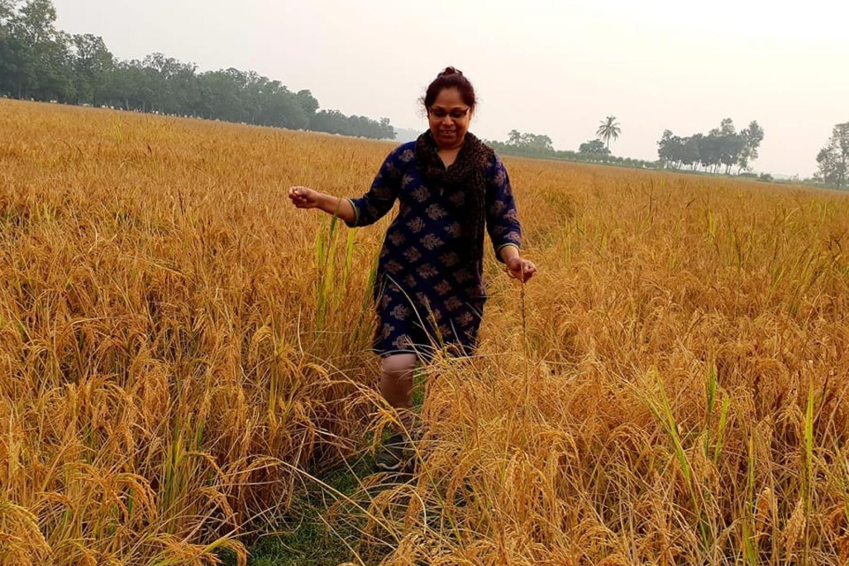 Woman walking through a field with trees visible in the distance. 