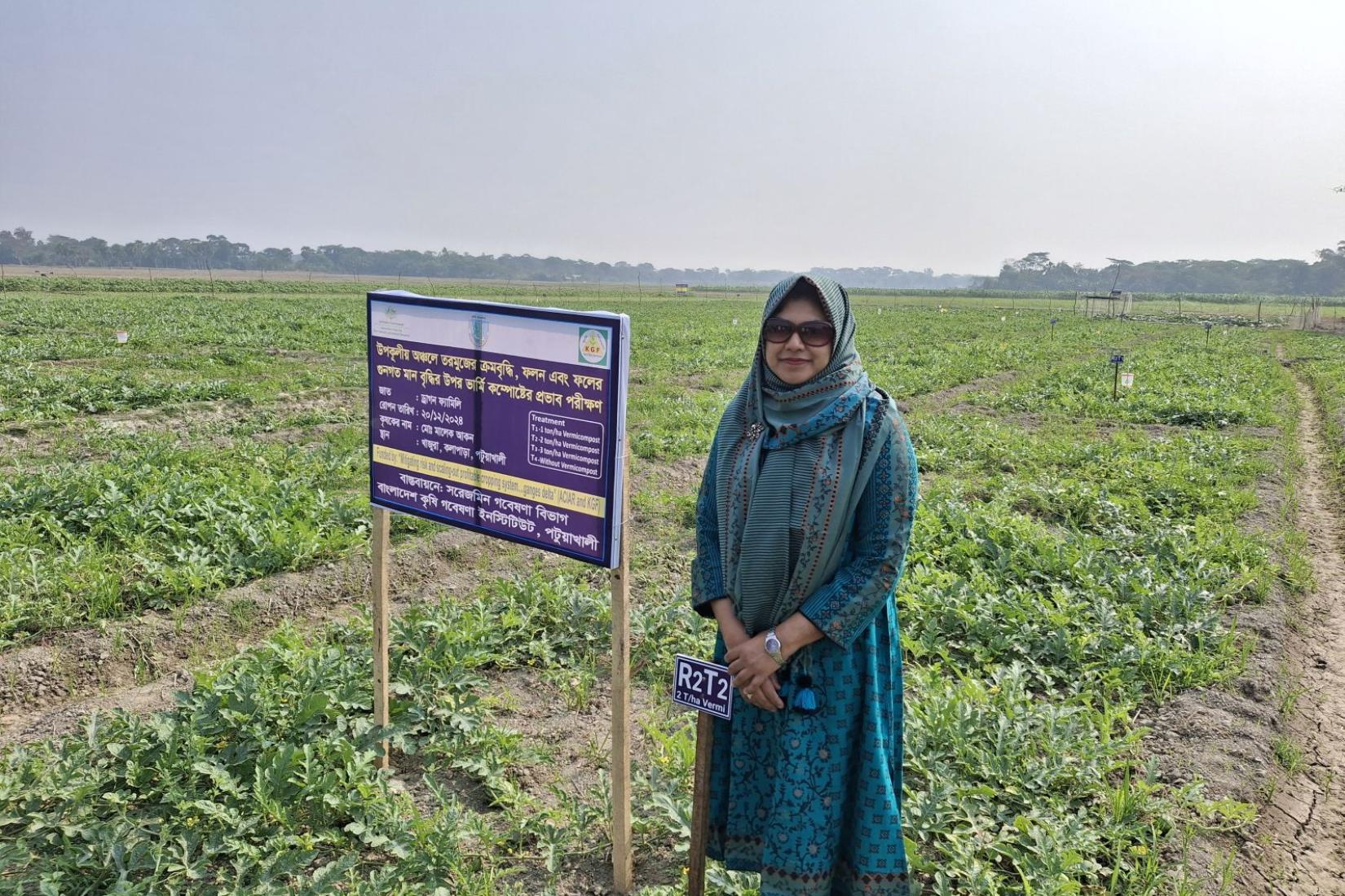 Taslima wearing a green headscarf and sunglasses standing in front of a crop field beside a research signboard. 