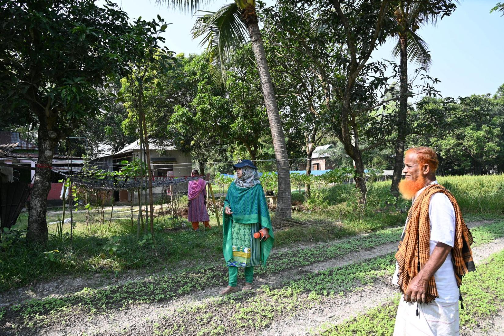 Three people standing outdoors near a small garden and trees in a rural village, with houses visible in the background.