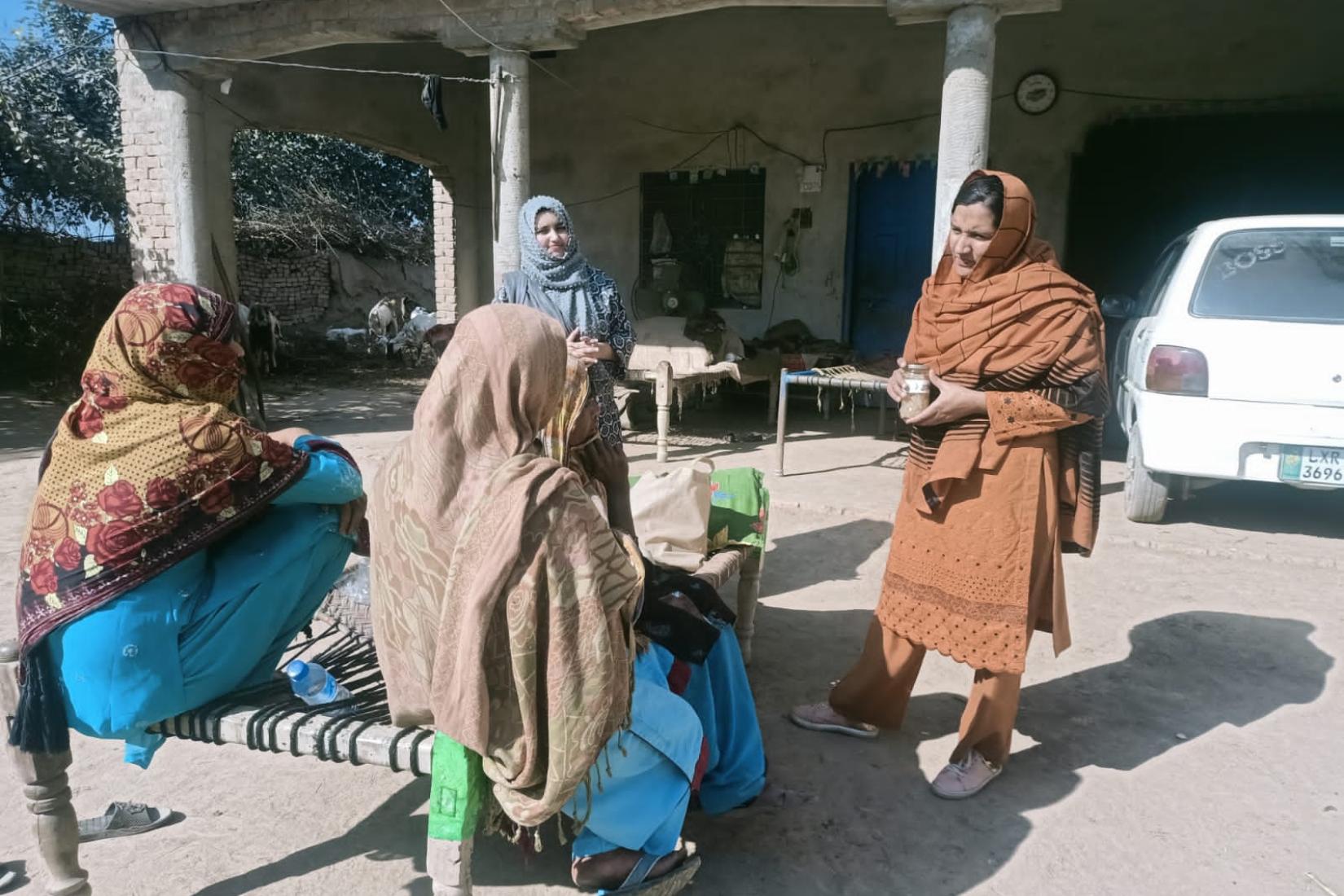 Four women gathered outdoors, talking near a house, with two seated on a woven bench and others standing nearby.