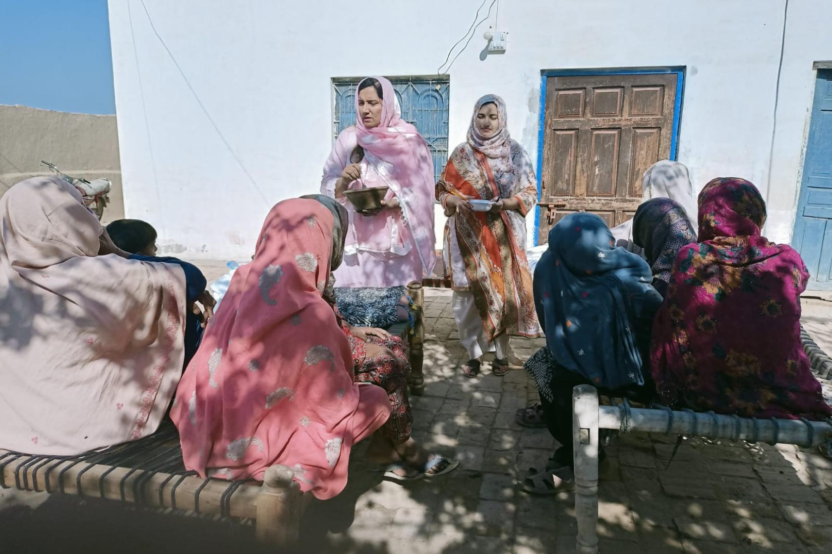 Women gathered outdoors in a courtyard, listening to 2 women standing and speaking.