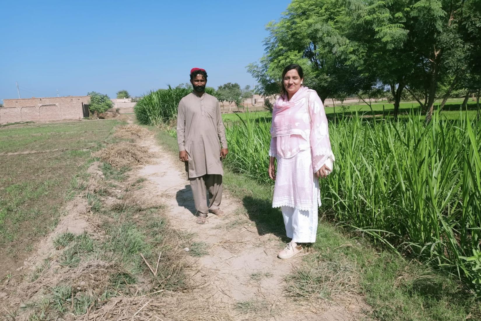Two people walking along a dirt path beside tall green crops and trees in a rural landscape.