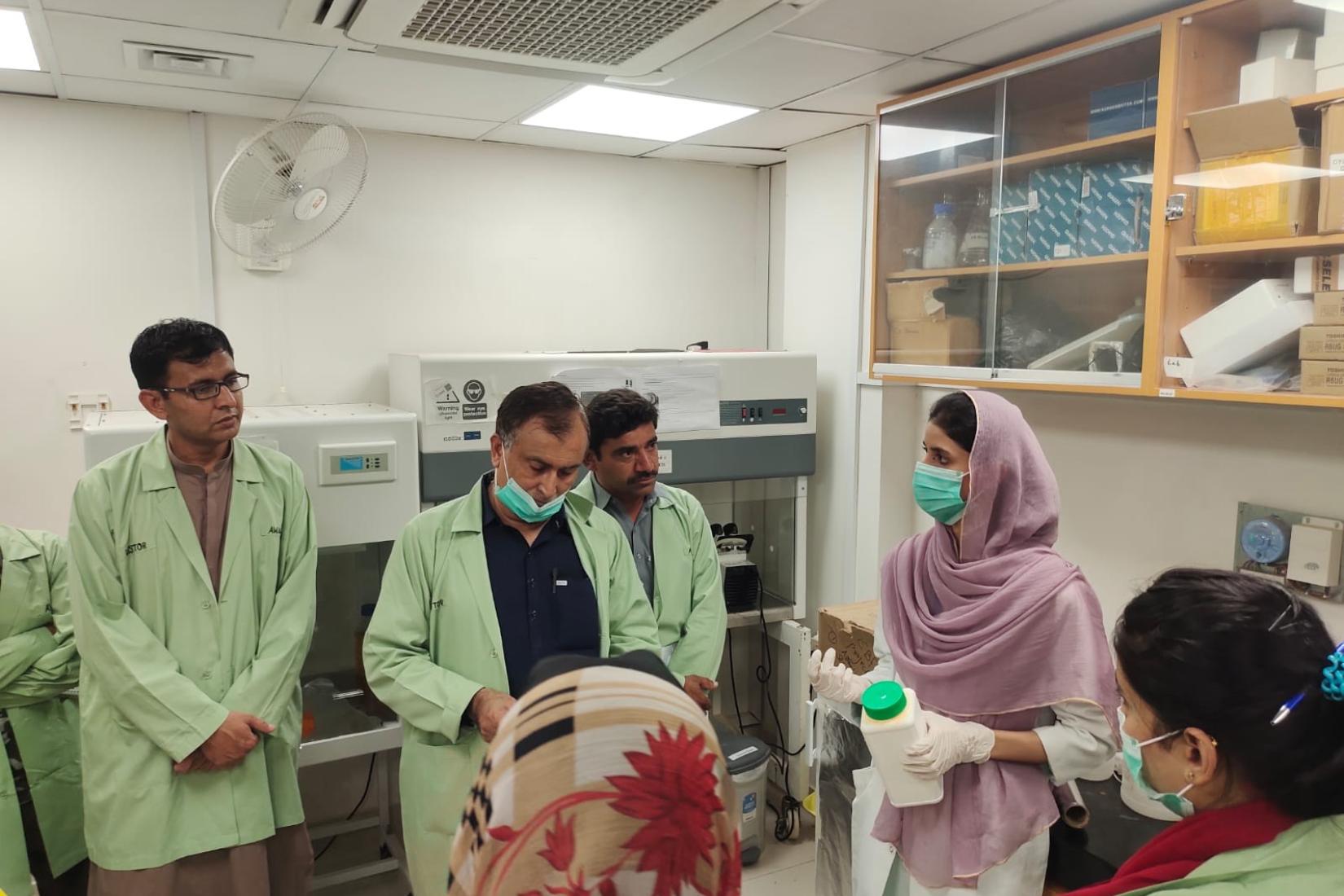 A group of people in lab coats gathered inside a laboratory, discussing and examining a container among scientific equipment and workbenches