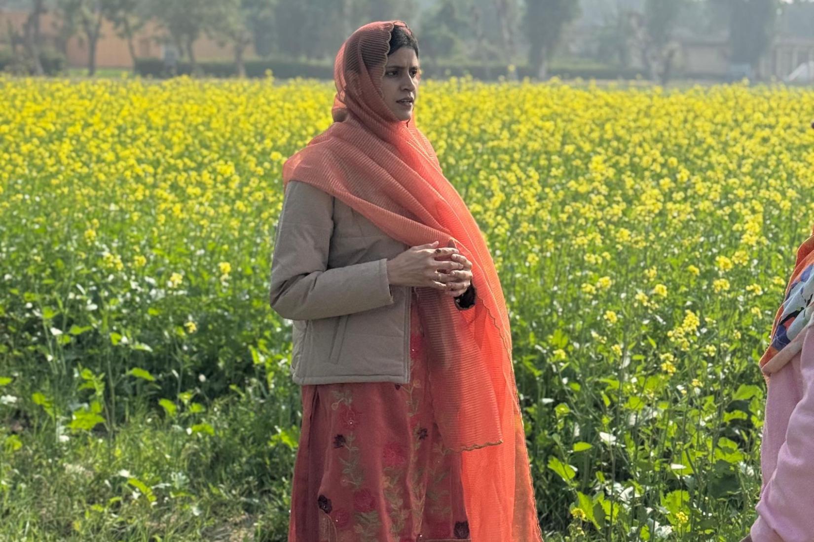 A woman standing in a field of yellow flowering crops, with trees visible in the background.