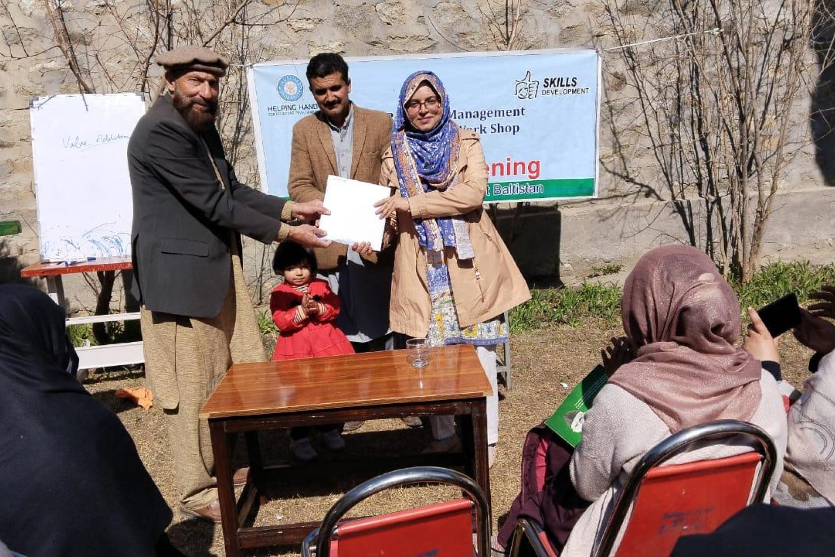 An outdoor certificate presentation with facilitators handing a document to a participant while attendees sit and watch.