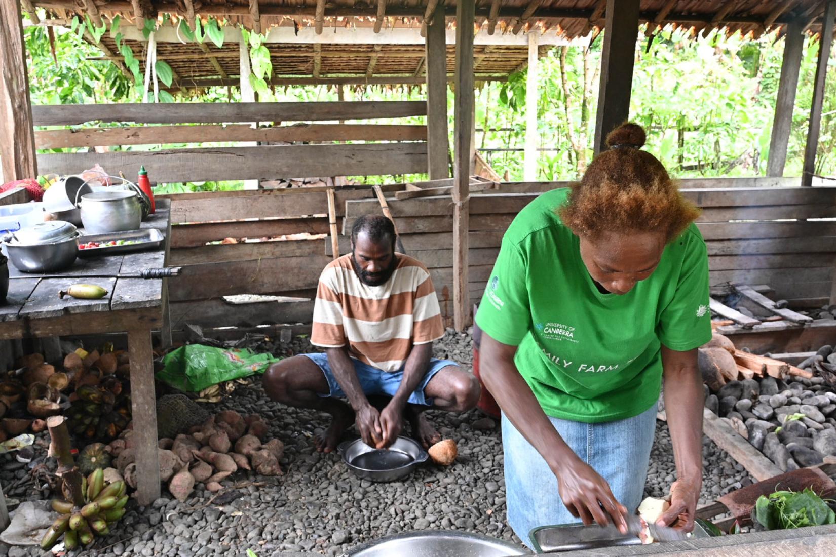 Two people prepare food together in an open kitchen shelter with cooking pots and root crops