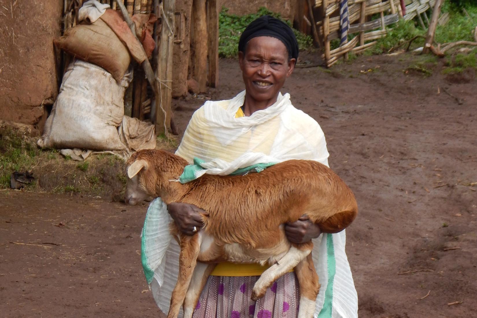 A smallholder farmer holding a young goat beside another goat in a village area.