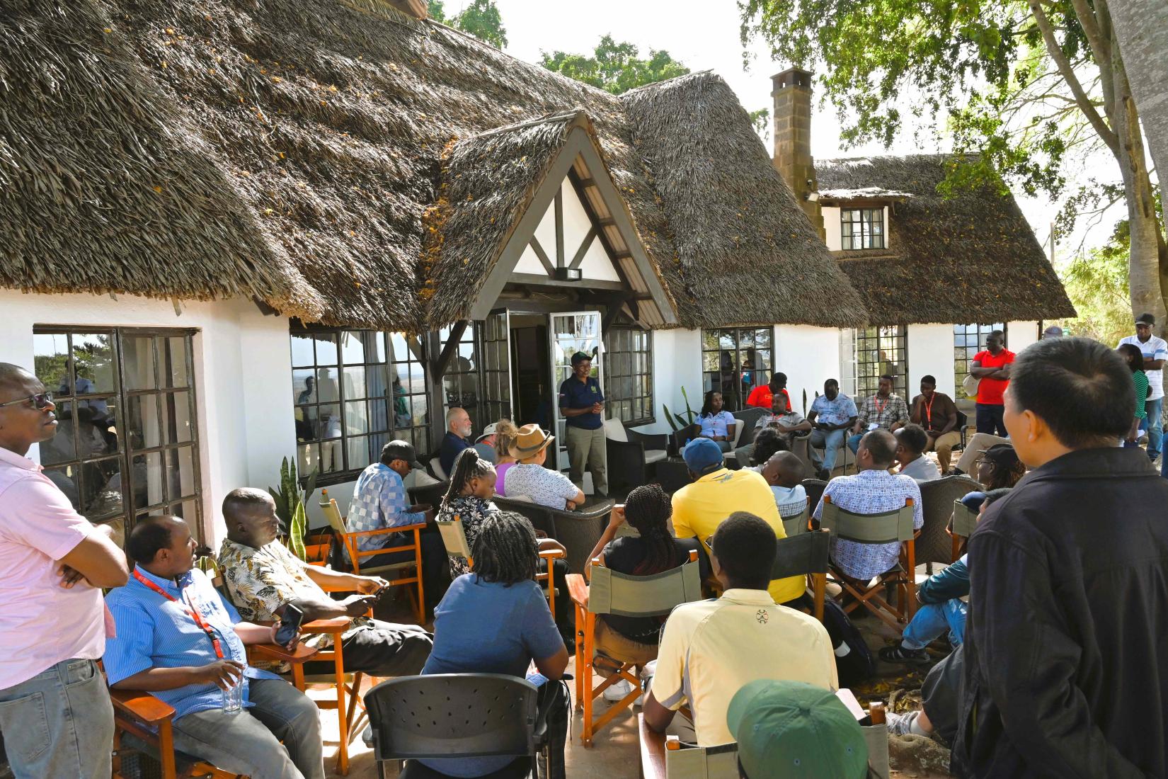 articipants seated outdoors, listening to a speaker at a lodge-style building.