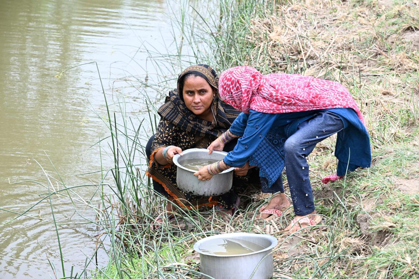 Two people collecting water from a canal using metal pots.