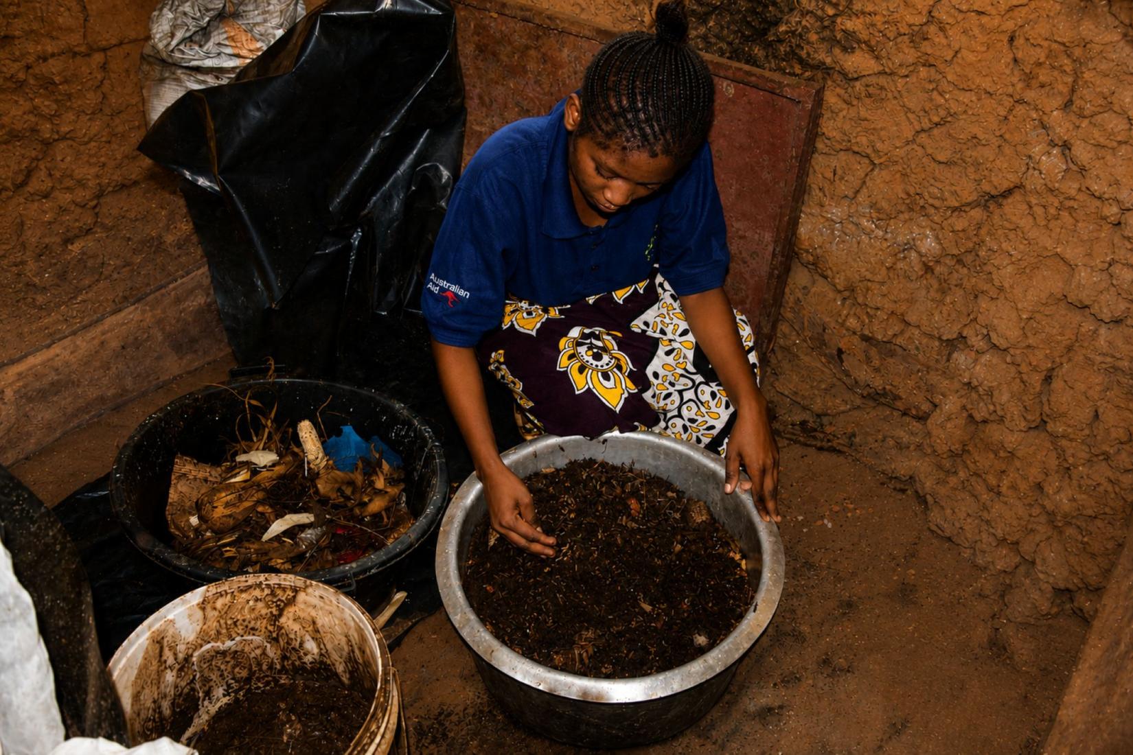 Person sorting organic material and insect larvae in a large basin inside a mud-brick structure.