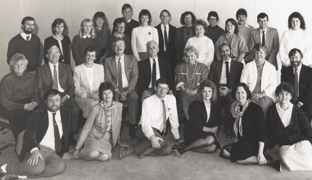 A black-and-white group photo of about 30 men and women in business attire. There are about three rows of people: the back row is standing, the centre row is sitting in chairs and the front row is sitting on the floor. 