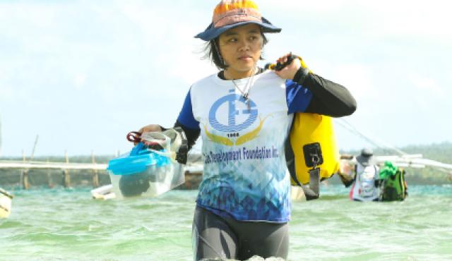 A woman stands in shallow ocean water. She is carrying bags and wears a white shirt and an orange hat.