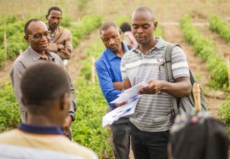 A group of men in a field, with one holding papers, sharing information with the others.