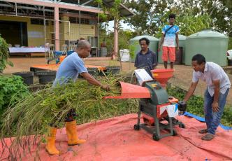Farmer feeds cut grass into a small chopper machine while others observe beside water tanks and a rural building
