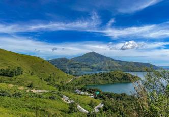 Green hills and a lake surrounded by mountains under a bright blue sky.