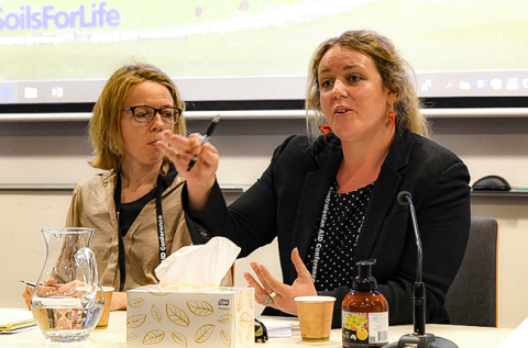 Two women sitting at a table. One woman is speaking to the audience with her hand raised. There are microphones on the table and a box of tissues and glasses of water. 