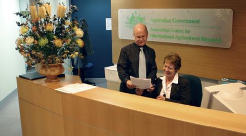A man standing next to a woman behind a reception desk. The woman is sitting next to a computer. The man standing next to her holding out some paper. There is a large vase of flowers on the reception bench. 