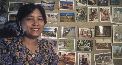 A woman with short black hair, wearing a flowered top, stands in front of a corkboard that has a lot of photographs stuck to it with thumbtacks.