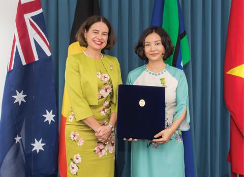 Two women stand on a stage. One is wearing a green dress and jacket with a flower pattern, and the other is wearing a light blue and white dress. The one in the green dress has presented the other with an award, which she is holding. Behind them are stage curtains and to the left the Australian flag. 