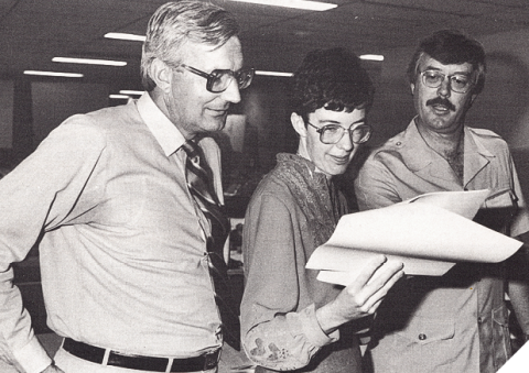 Black-and-white photo of two men with a woman standing in the middle of them holding and looking down at pieces of paper. The woman and one of the men are wearing glasses