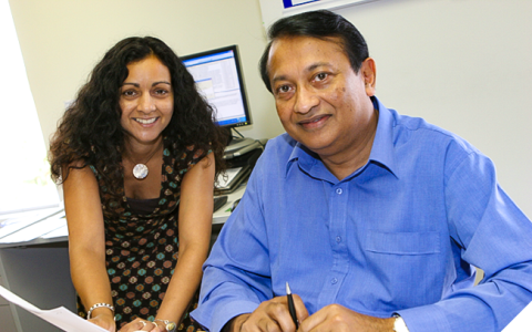 A man and a woman sitting next to each other at a desk. He has black hair and is wearing a blue-collared shirt. She has black curly hair past her shoulders and is wearing a printed black dress. They are smiling