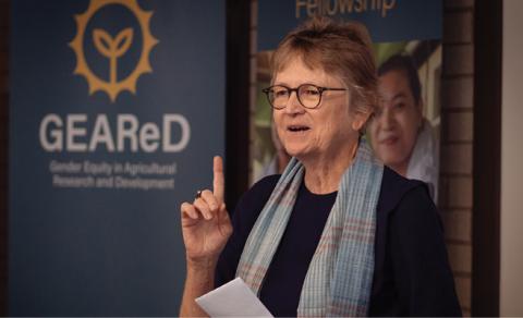 A photograph of Meryl Williams. She wears glasses and a scarf is talking with her hand raised. 