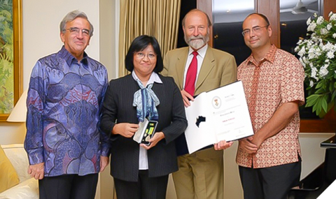 Three men standing next to a woman. She was wearing a black suit and holding an award. The man with a red tie standing next to her is holding an open book. They are all smiling. 