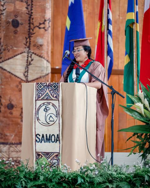 Woman standing at a lecturn on a stage in graduation attire