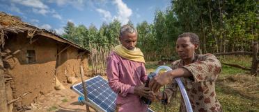 A man and woman setting up a solar panel next to a mud hut