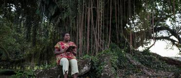 A woman sitting in a forest, with a smart tablet in her hands, looking into the distance.