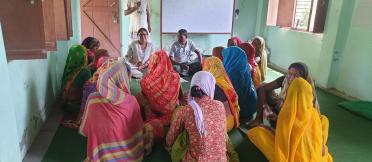 Group of women in colourful clothing seated in a circle indoors, listening to two facilitators, with a whiteboard behind them. 