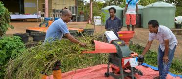 Farmer feeds cut grass into a small chopper machine while others observe beside water tanks and a rural building