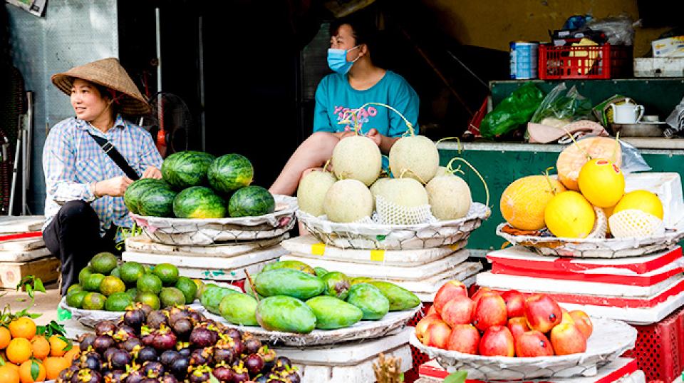 a variety of fruits in baskets, and two women sitting in the background. one of the women wears a large brimed traditional hat