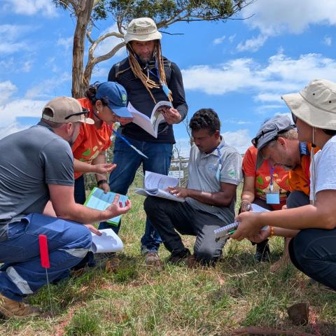 A group of people sit together in a field in discussion holding paper and pens.